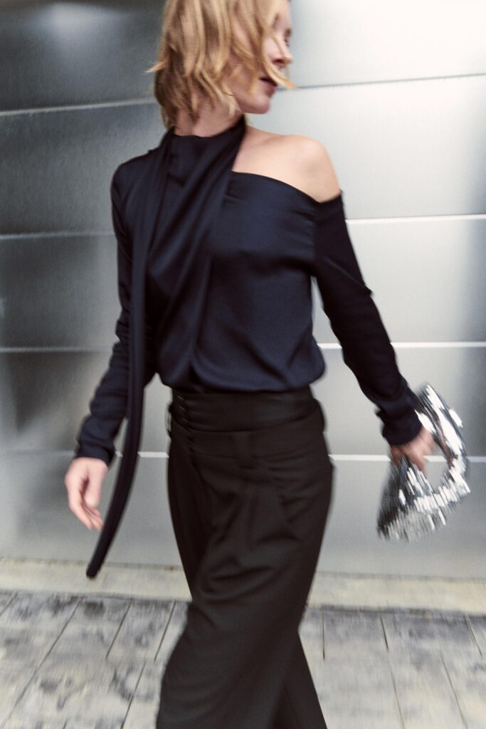 A fitted black silk off-shoulder top with long sleeves paired with tailored black pants featuring an asymmetric design.