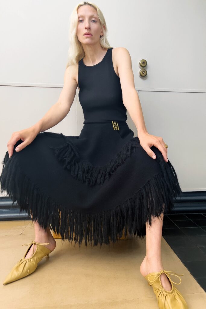 A fitted black sleeveless dress with a high neckline and fringed asymmetric detail.