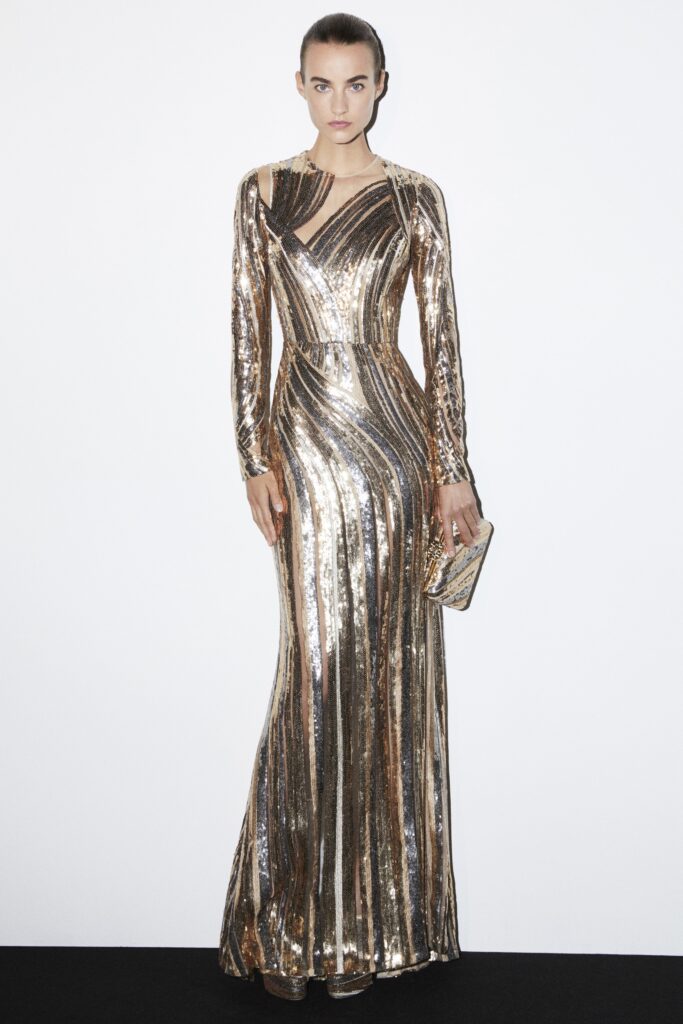 A fitted gold sequin gown with long sleeves and a high neckline featuring pleated details.