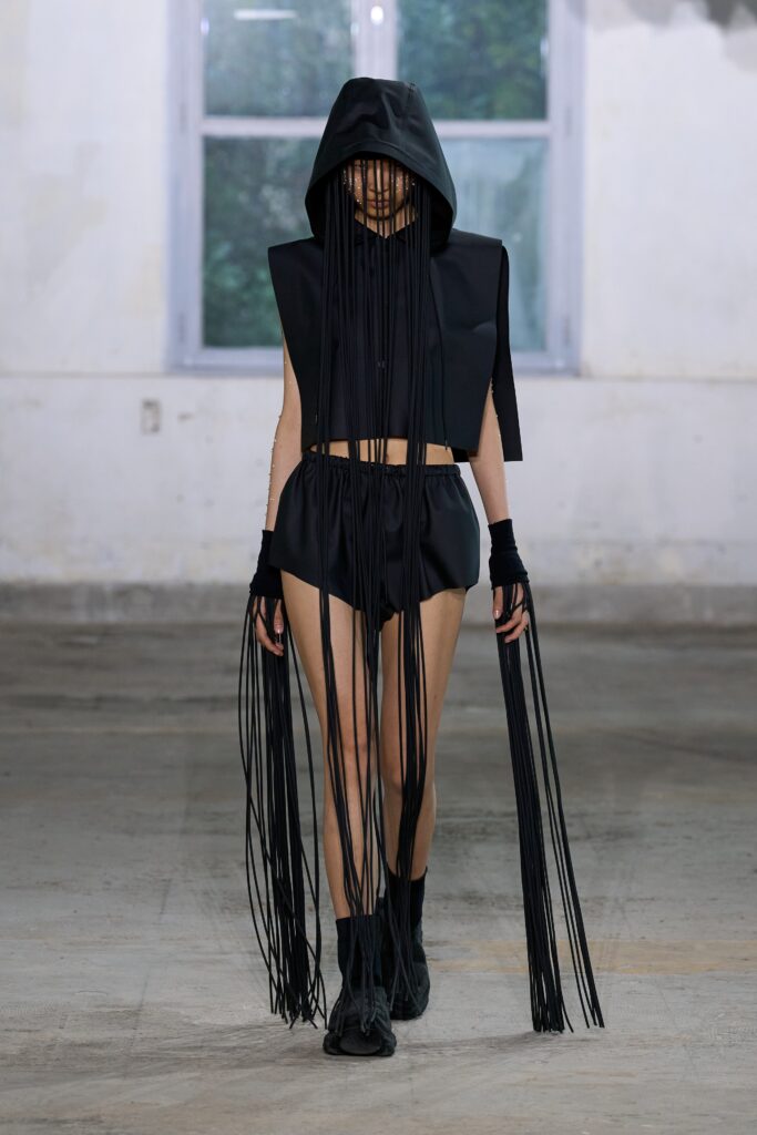 A fitted black sleeveless hooded top paired with fringed black shorts.