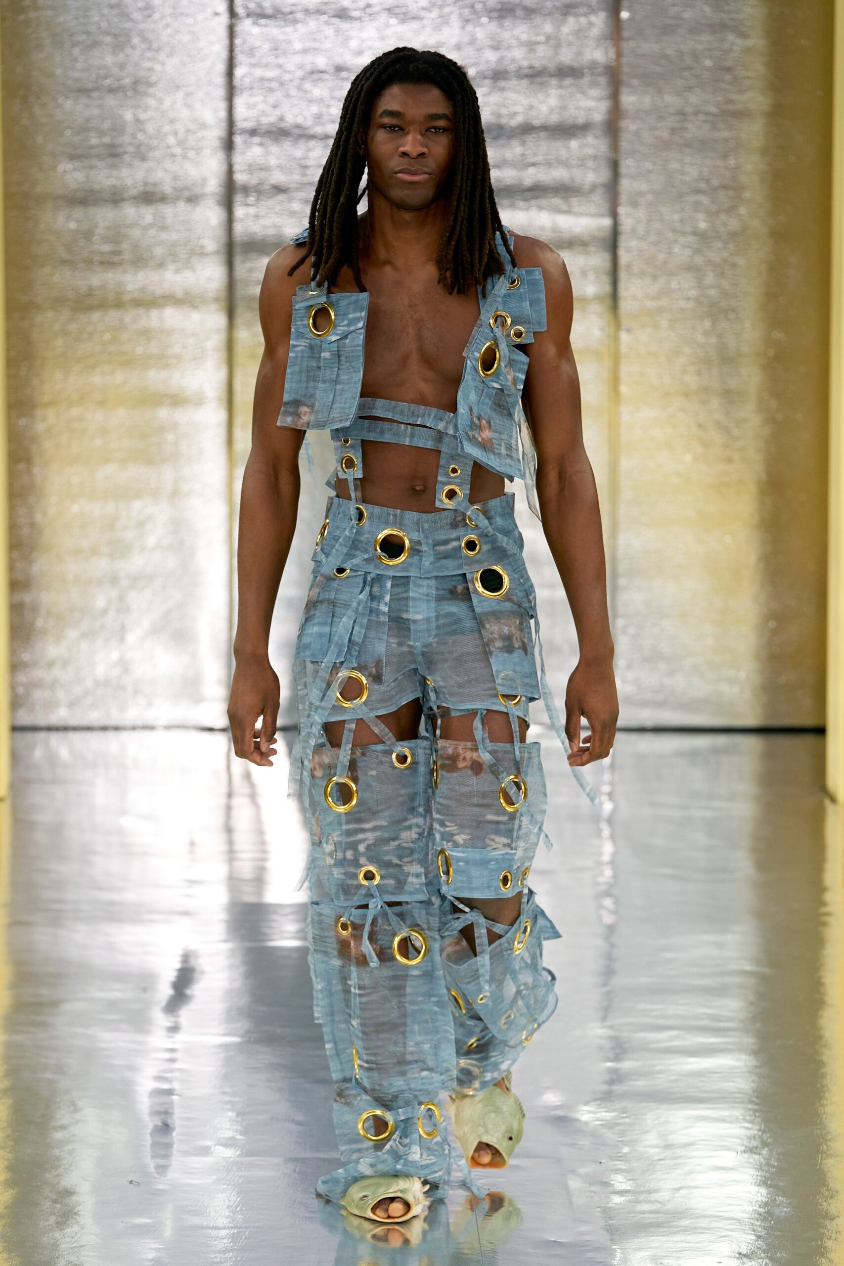 A fitted blue denim sleeveless jumpsuit with a V-neckline and asymmetric cutouts.