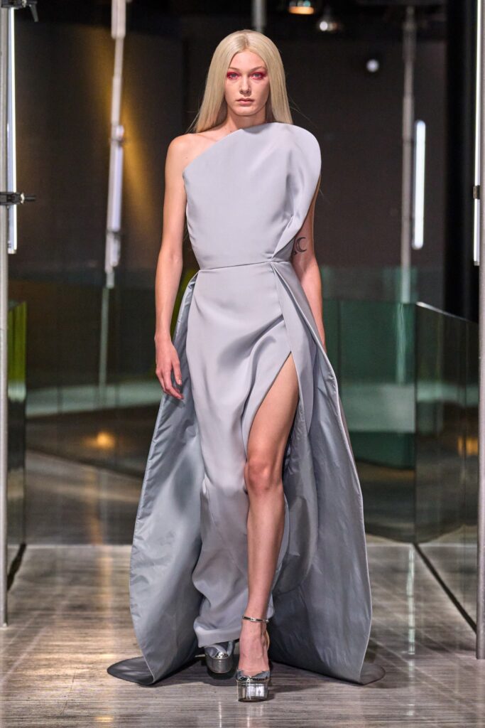 A fitted gray silk dress with a sleeveless design, high-neck, and an asymmetric silhouette featuring a slit.