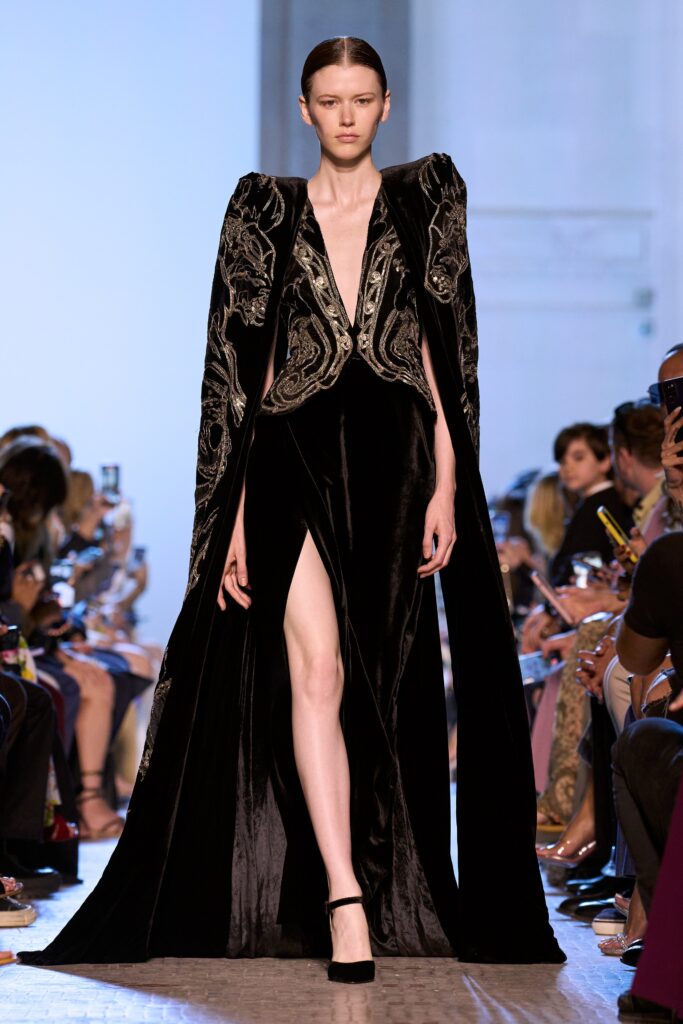 A fitted black velvet sleeveless dress with a V-neckline, slit, and an embroidered cape.