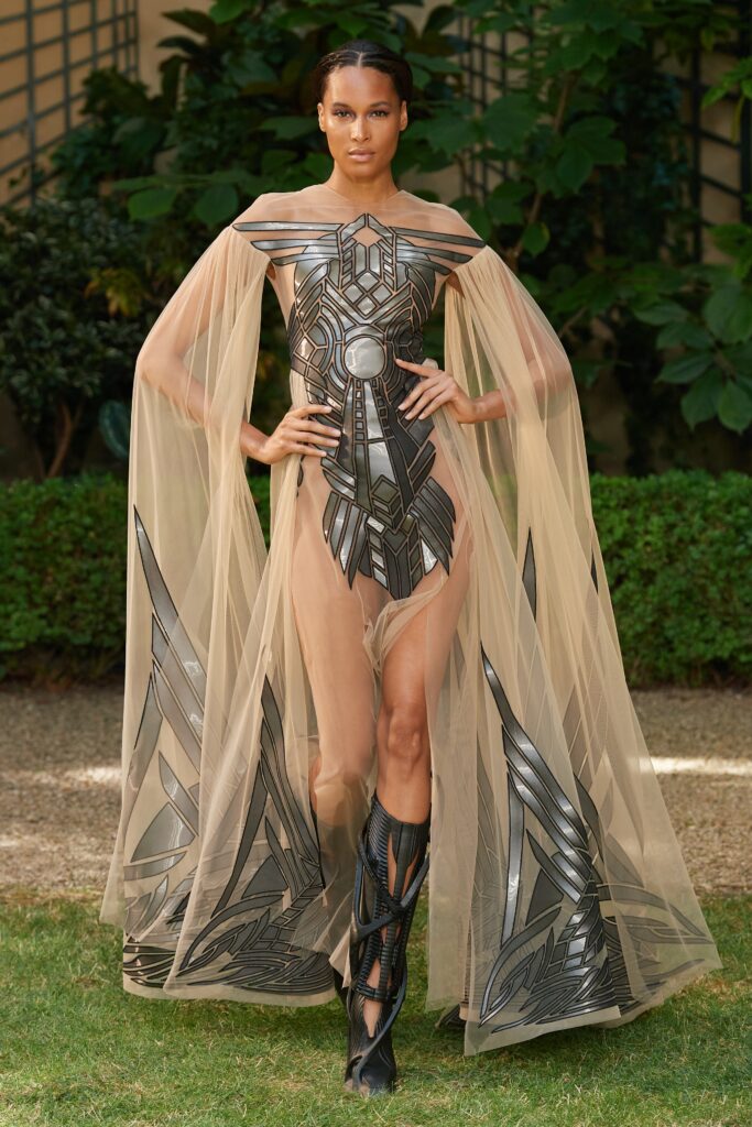 A fitted beige sheer dress with an off-shoulder design and a layered metallic cape.