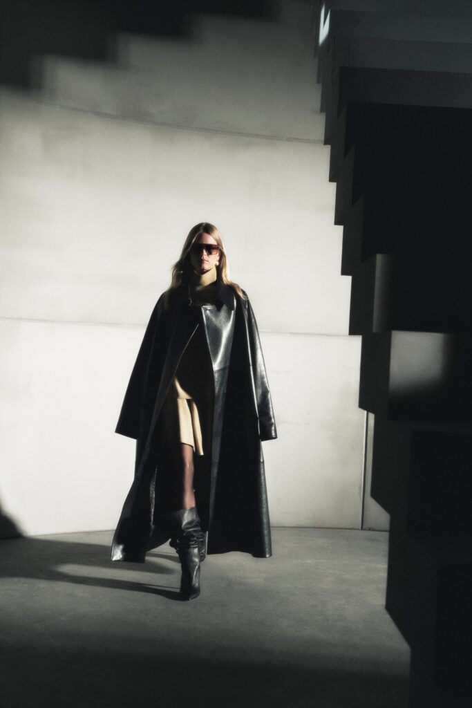 A fitted black leather coat worn over a sleeveless beige dress with an asymmetric design.