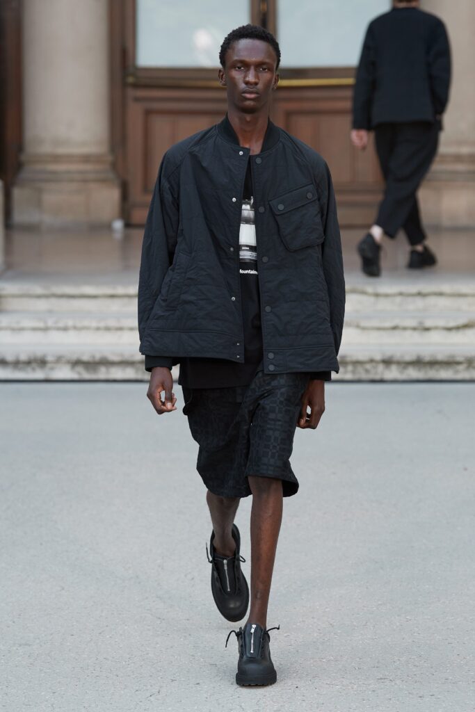 A relaxed black cotton jacket with short sleeves paired with black shorts featuring a zippered detail.