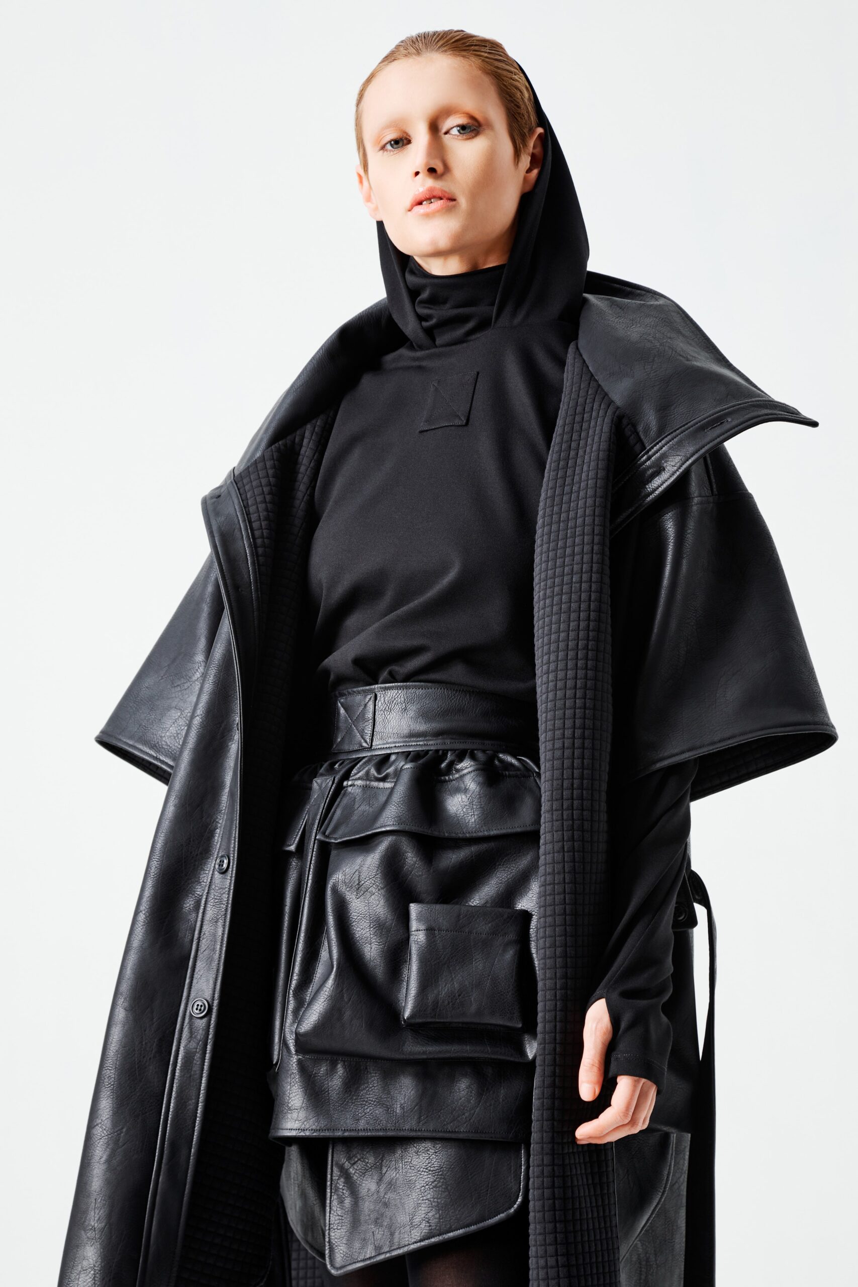 This ensemble features a fitted black leather coat layered over a sleek top, both designed with a contemporary edge. The long sleeves and hood add a functional yet stylish element, while the layered construction creates a dynamic silhouette that plays with proportion and depth.
