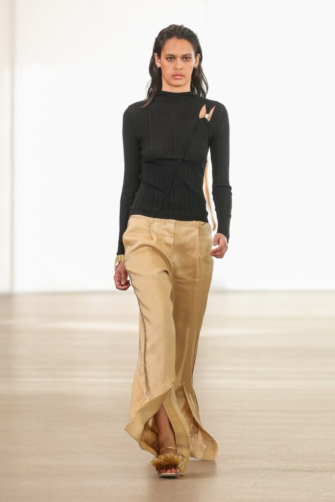 A fitted black long-sleeve top with a V-neck and asymmetric design paired with beige straight-leg pants.