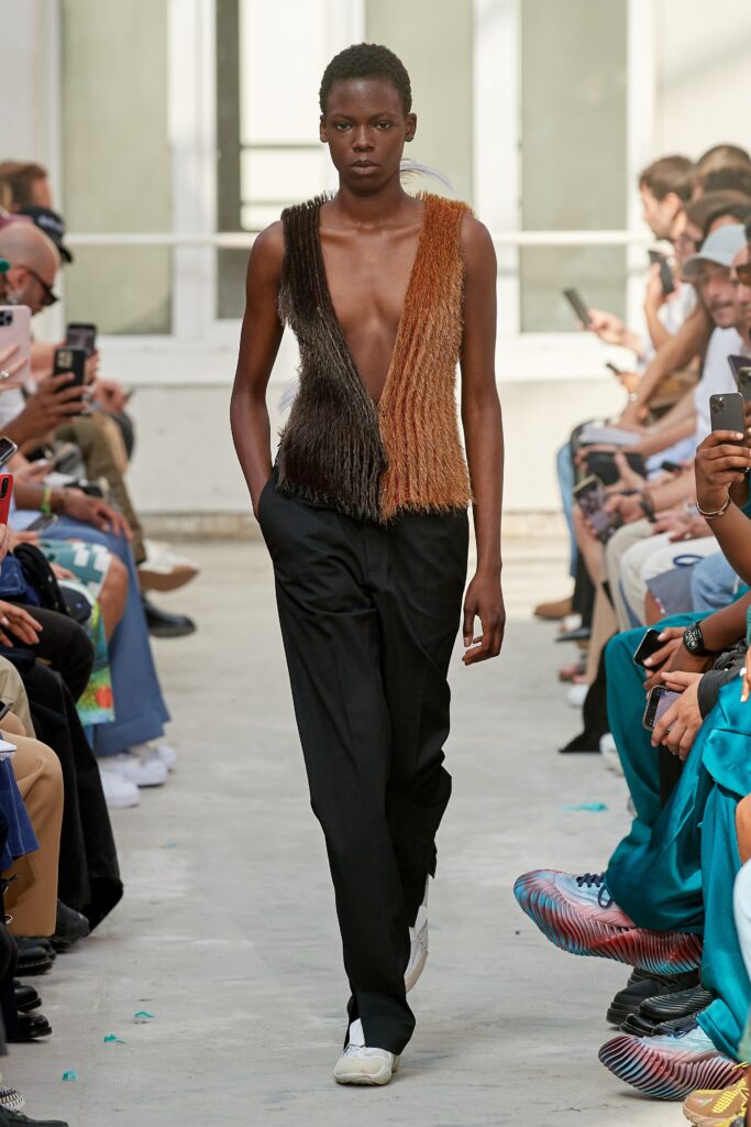 A fitted brown leather sleeveless top with a V-neck and asymmetric design paired with black pants.