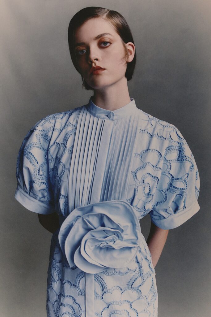 A fitted blue cotton dress with short sleeves, a collared neckline, and pleated embroidered details.