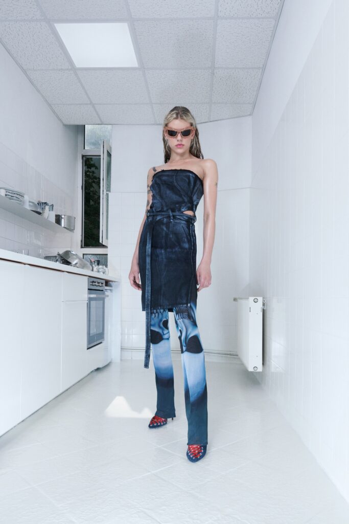A fitted black sleeveless dress with asymmetric design and fringed detail paired with blue pants.