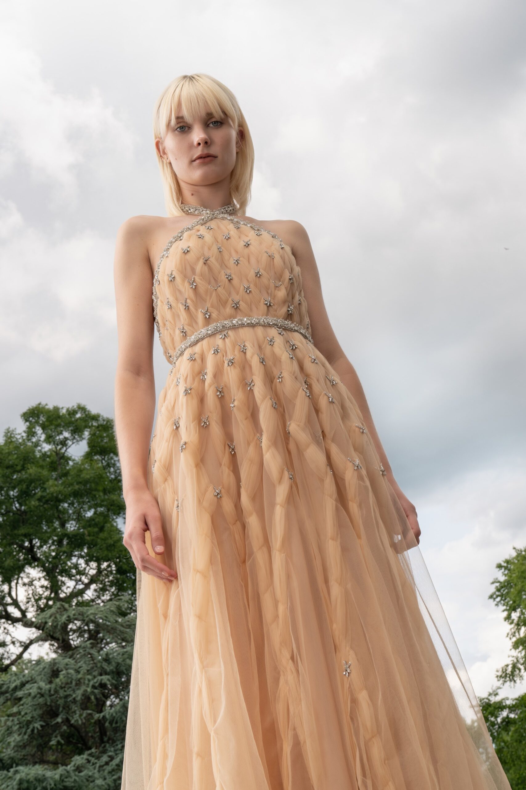 A fitted beige tulle sleeveless gown with a high neckline and layered design.