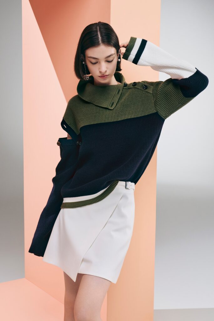 A relaxed navy and green long-sleeve sweater with an asymmetric design paired with a white skirt.