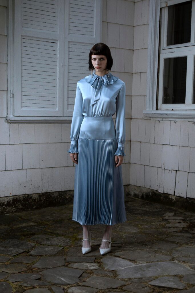 A fitted blue silk dress with long sleeves, a high neckline, and pleated detailing.