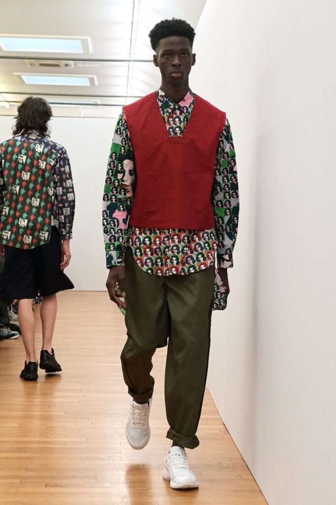 A relaxed red cotton vest worn over a long-sleeve printed shirt.