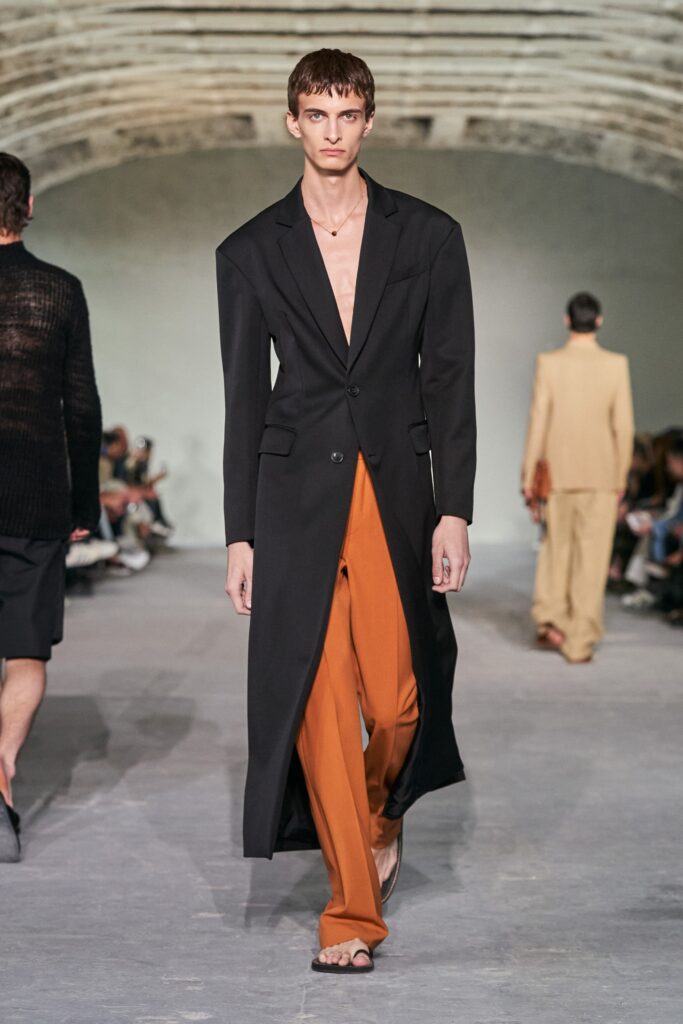 A relaxed black coat paired with orange pants featuring an asymmetric design.