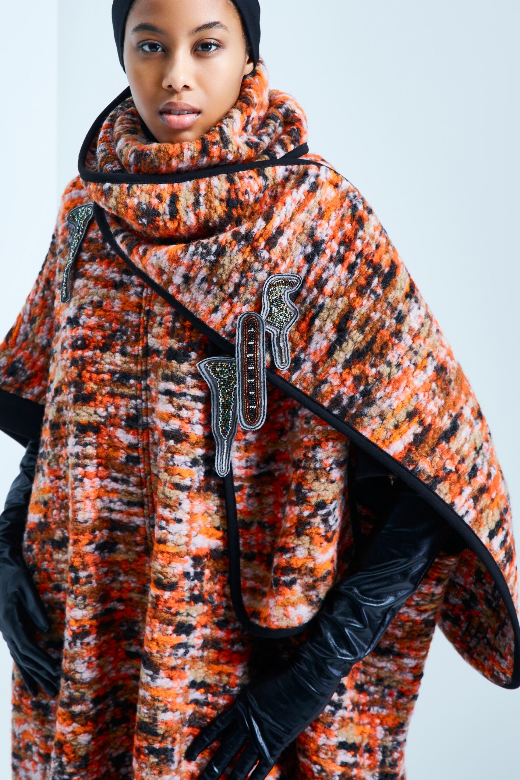 This relaxed cape showcases a vibrant interplay of orange, black, and white hues, creating a striking visual texture. The high-neck design adds a contemporary touch, while the layered silhouette offers a sense of movement and depth.