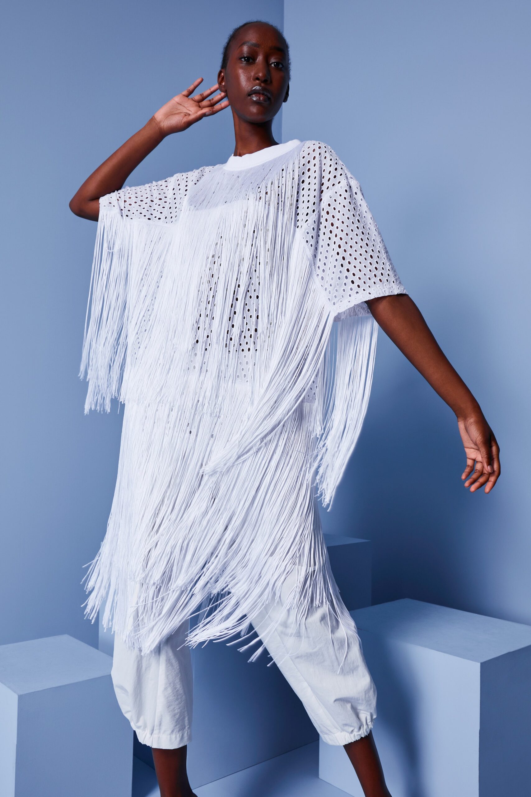 This oversized top features an intricate fringe detail that adds dynamic movement to its silhouette. Crafted from lightweight cotton, it showcases a relaxed fit with short sleeves and a collared design, creating a striking visual contrast against the soft background.