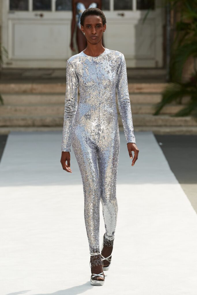 A fitted silver sequin catsuit with long sleeves and a high neckline featuring a zippered detail.