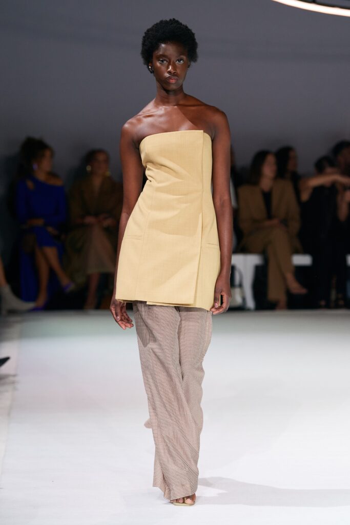 A fitted beige sleeveless top with an asymmetric design paired with brown pants.