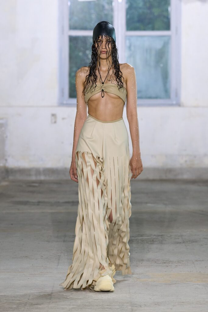 A fitted beige silk sleeveless dress with a high neckline and pleated asymmetric design.