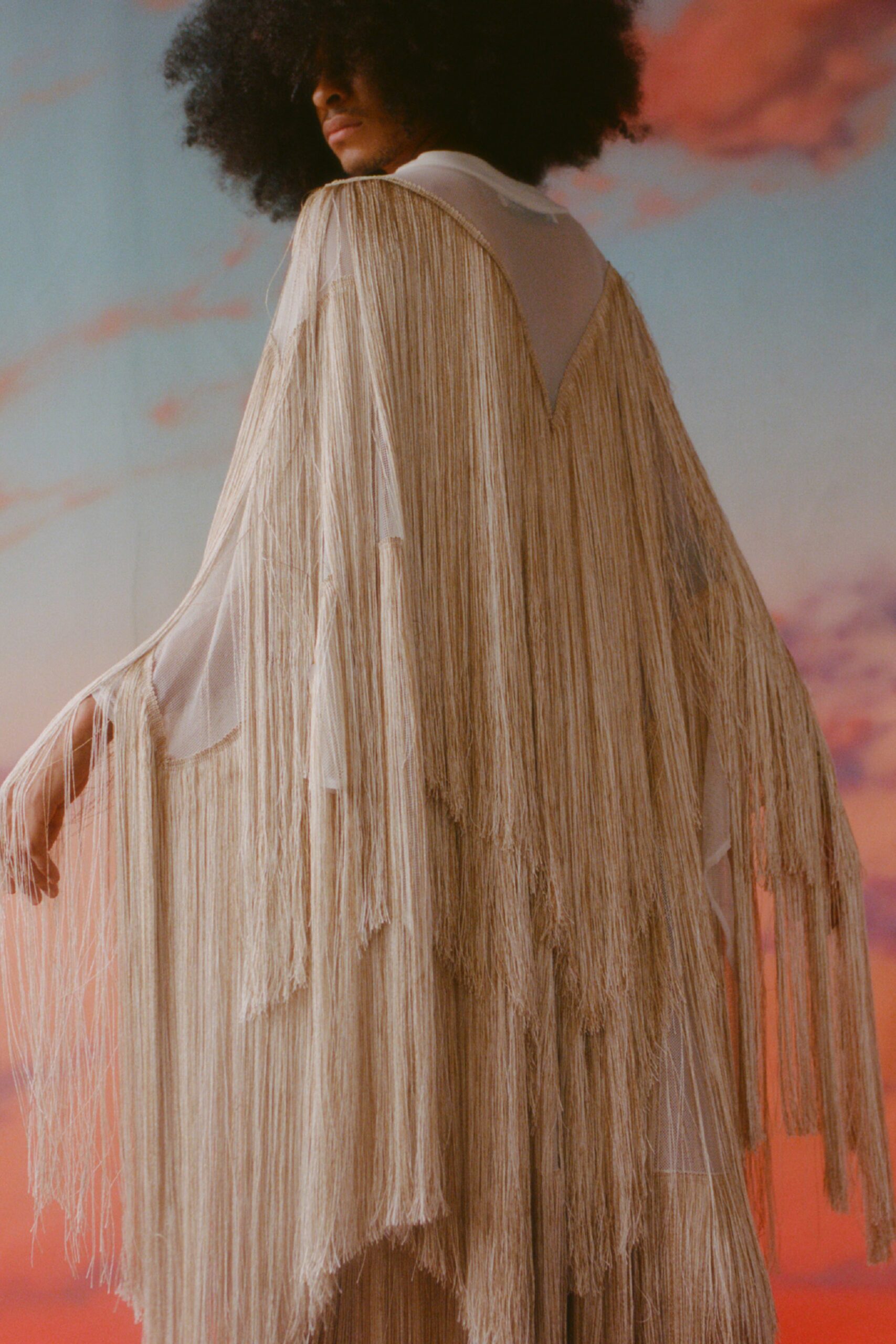 This relaxed cape in beige silk features an eye-catching fringed design that adds dynamic movement. The V-neckline enhances the flowing silhouette, creating a striking visual that captures attention with its layered texture.
