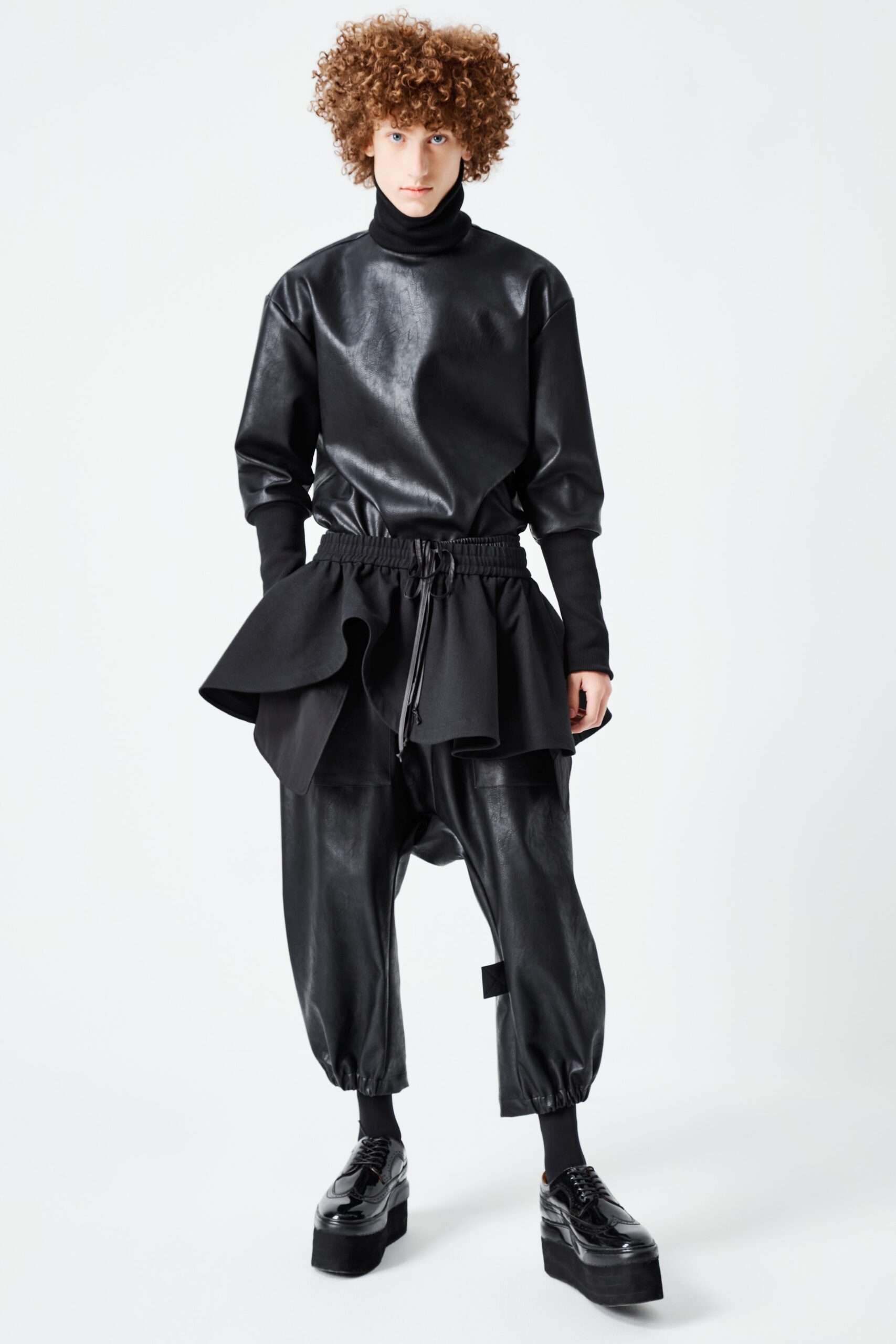 This ensemble features a relaxed black leather top with a high neck and long sleeves, complemented by pants that showcase an asymmetric layered design. The interplay of textures and silhouettes creates a dynamic visual interest, enhancing the overall modern aesthetic.