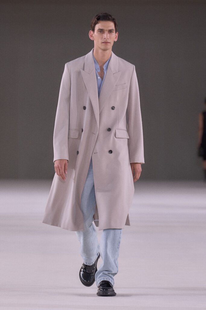 A relaxed beige coat layered over a collared blue shirt, featuring buttoned details.