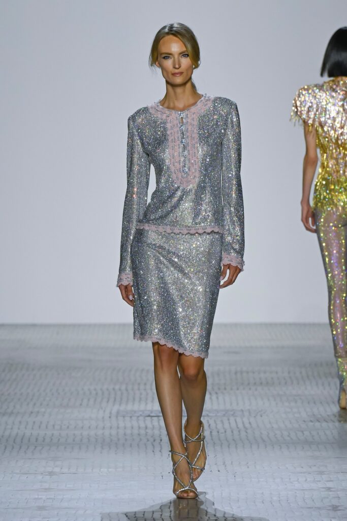A fitted silver sequin blouse with long sleeves and a collared neckline paired with an asymmetric skirt.