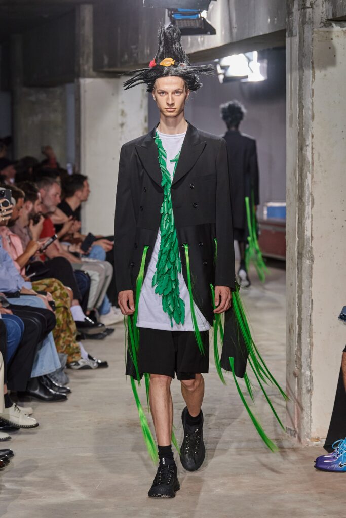 A relaxed black coat layered over a green fringed shirt, featuring long sleeves and a collared design.