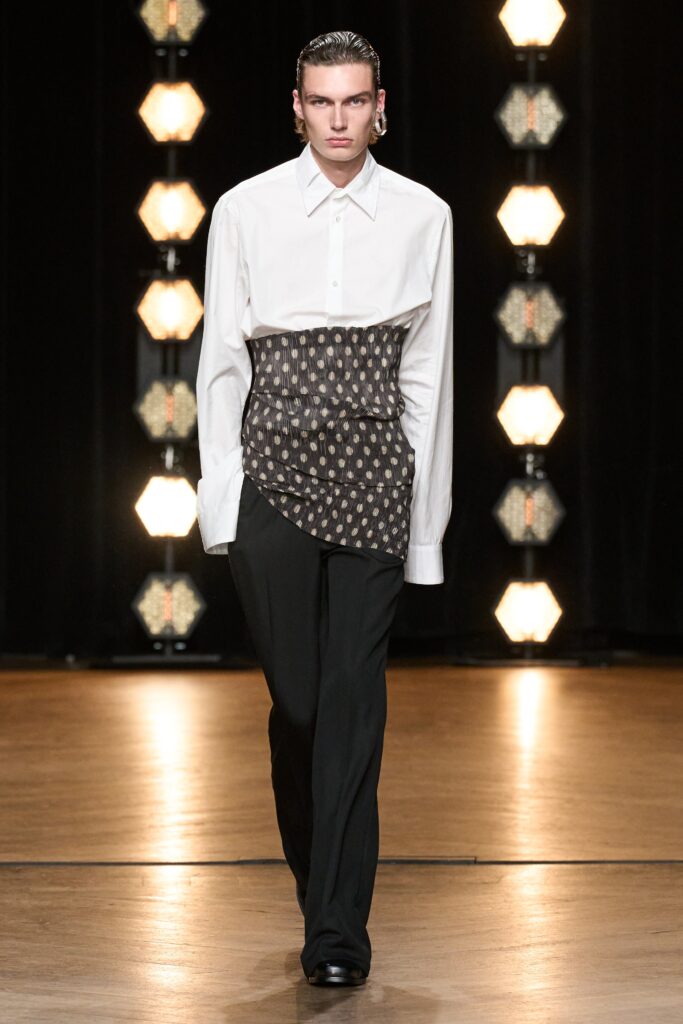A fitted white long-sleeve shirt with an asymmetric printed detail and black pants.