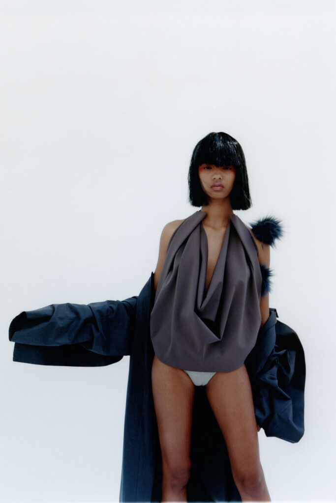 A relaxed black coat worn over a sleeveless gray draped top with a V-neckline.