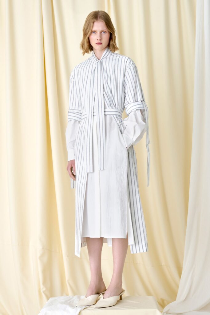 This dress features a relaxed silhouette with long sleeves and a collared neckline, accentuated by a belted waist that adds structure. The vertical stripes create a dynamic visual rhythm, enhancing the overall fluidity and movement of the garment.