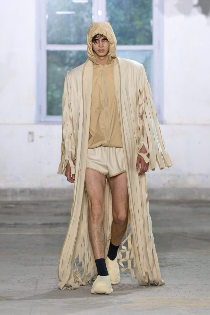 A relaxed beige coat with fringed details and a hood, paired with matching shorts.