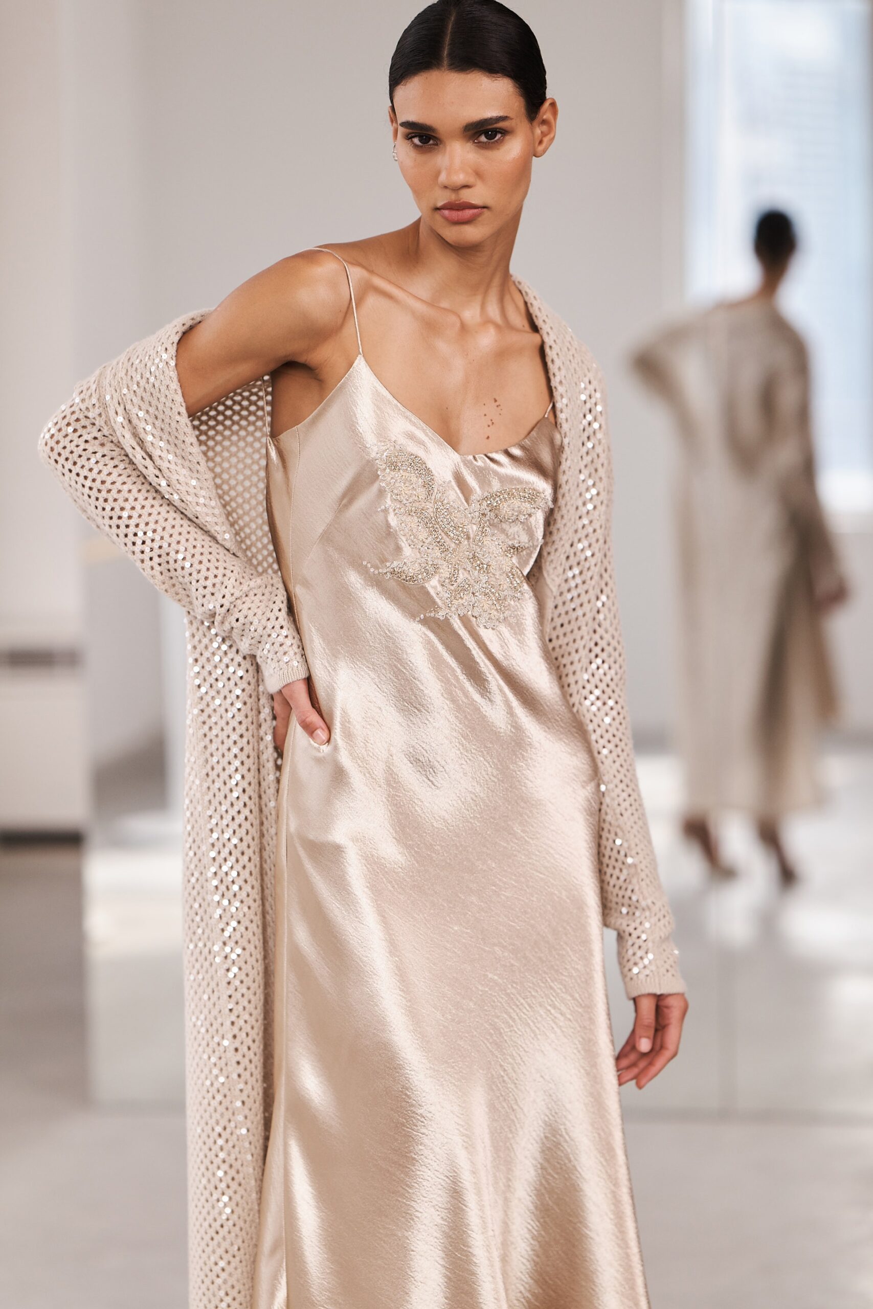 A fitted beige silk sleeveless dress with a V-neckline, layered with a knit cardigan.