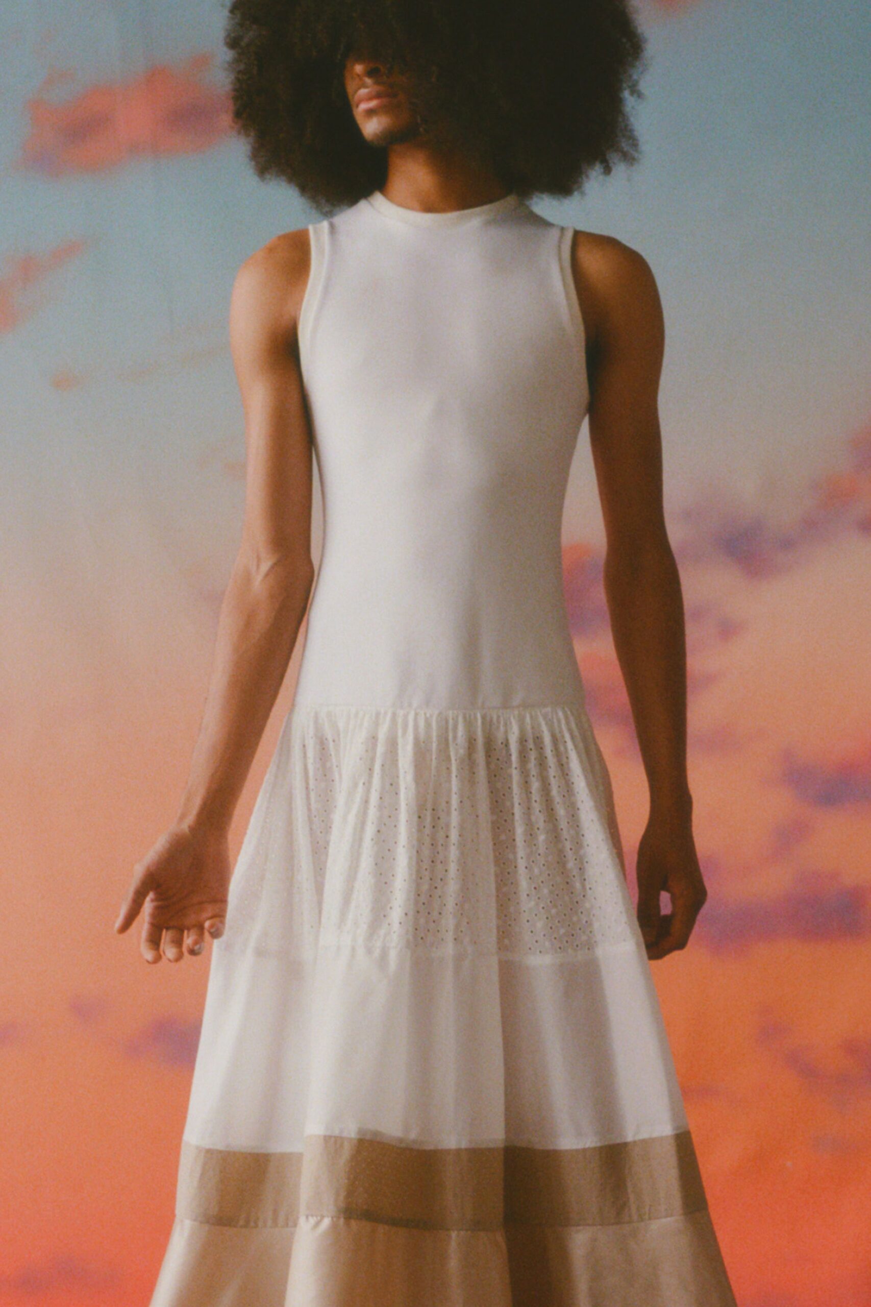 This sleeveless dress features a high-neck design that transitions into a flowing, tiered silhouette. The interplay of white and beige creates a soft visual contrast, enhancing its ethereal quality and movement.