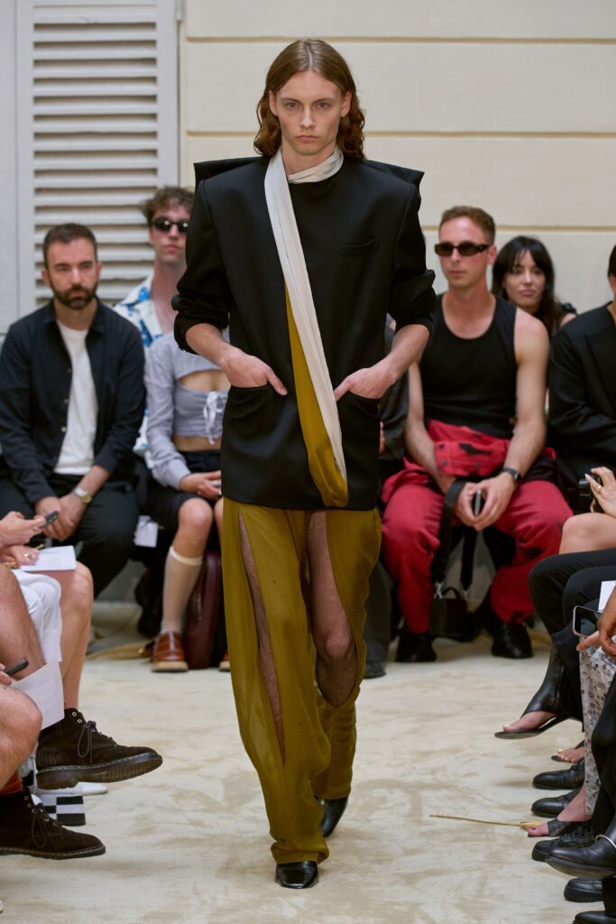 A relaxed black jacket with an asymmetric design paired with olive pants featuring a zippered detail.
