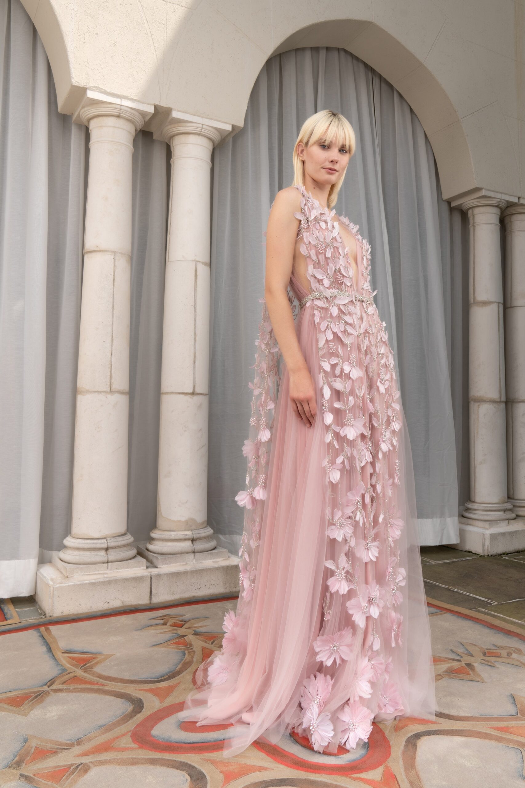 A fitted pink silk gown with a V-neckline, sleeveless design, and layered floral details.