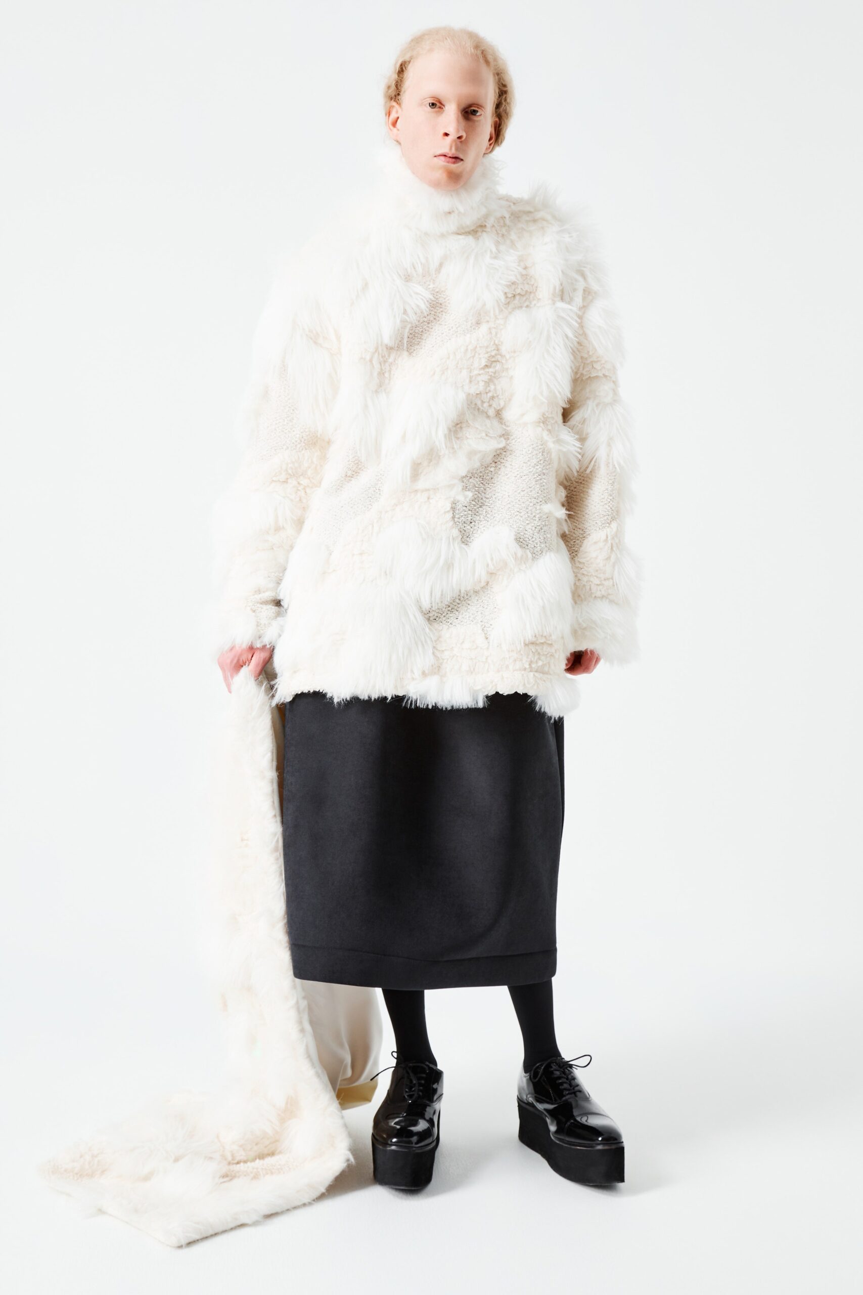 This relaxed coat features a high-neck design and long sleeves, crafted from plush faux-fur that adds depth and texture. Paired with a sleek black skirt, the ensemble showcases an intriguing interplay of layers and proportions, creating a striking visual narrative.