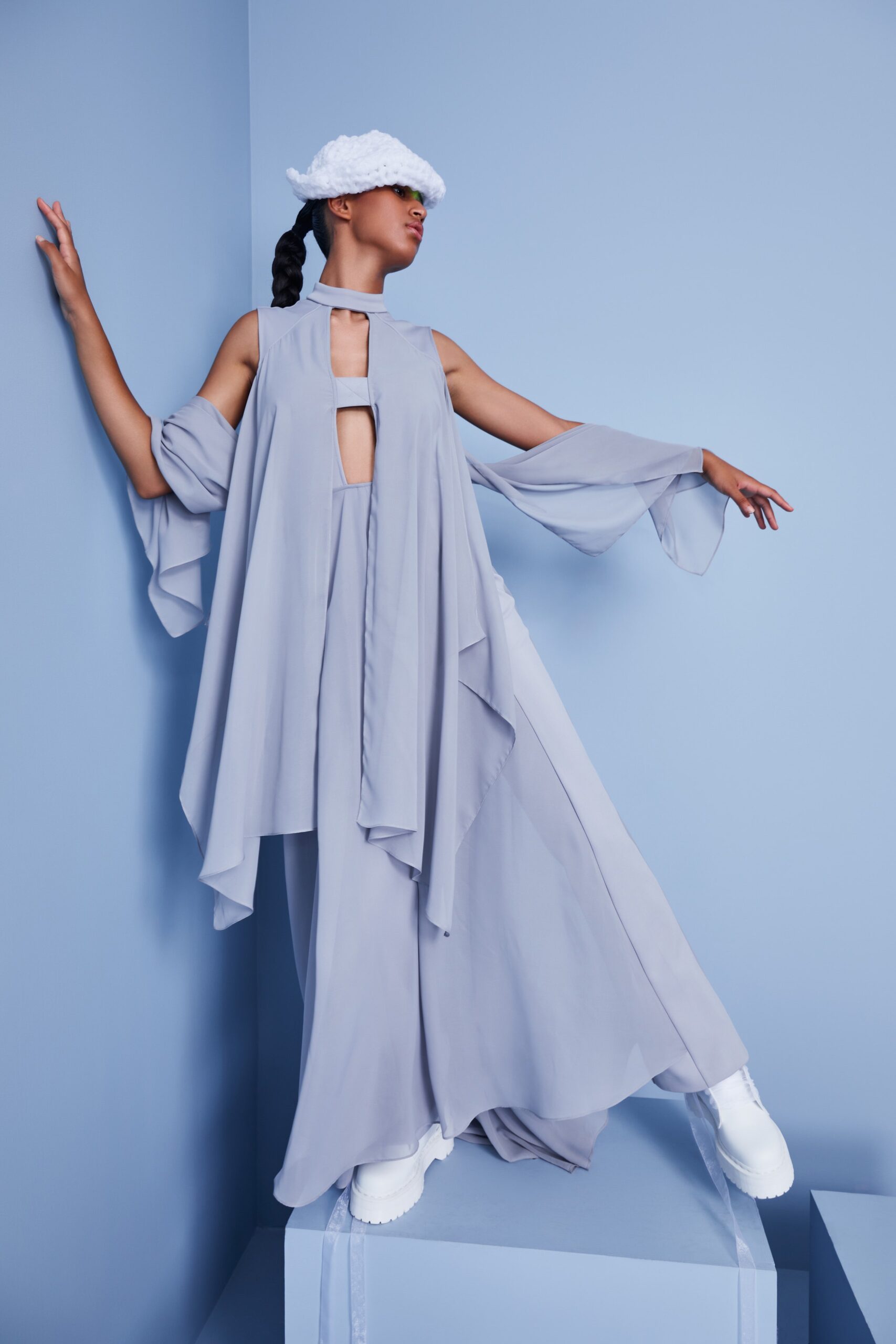 This relaxed dress features an airy silhouette crafted from soft chiffon, complemented by an asymmetric cape that adds fluidity to the overall look. The high-neck design enhances the ethereal quality, creating a striking visual interplay between structure and movement.