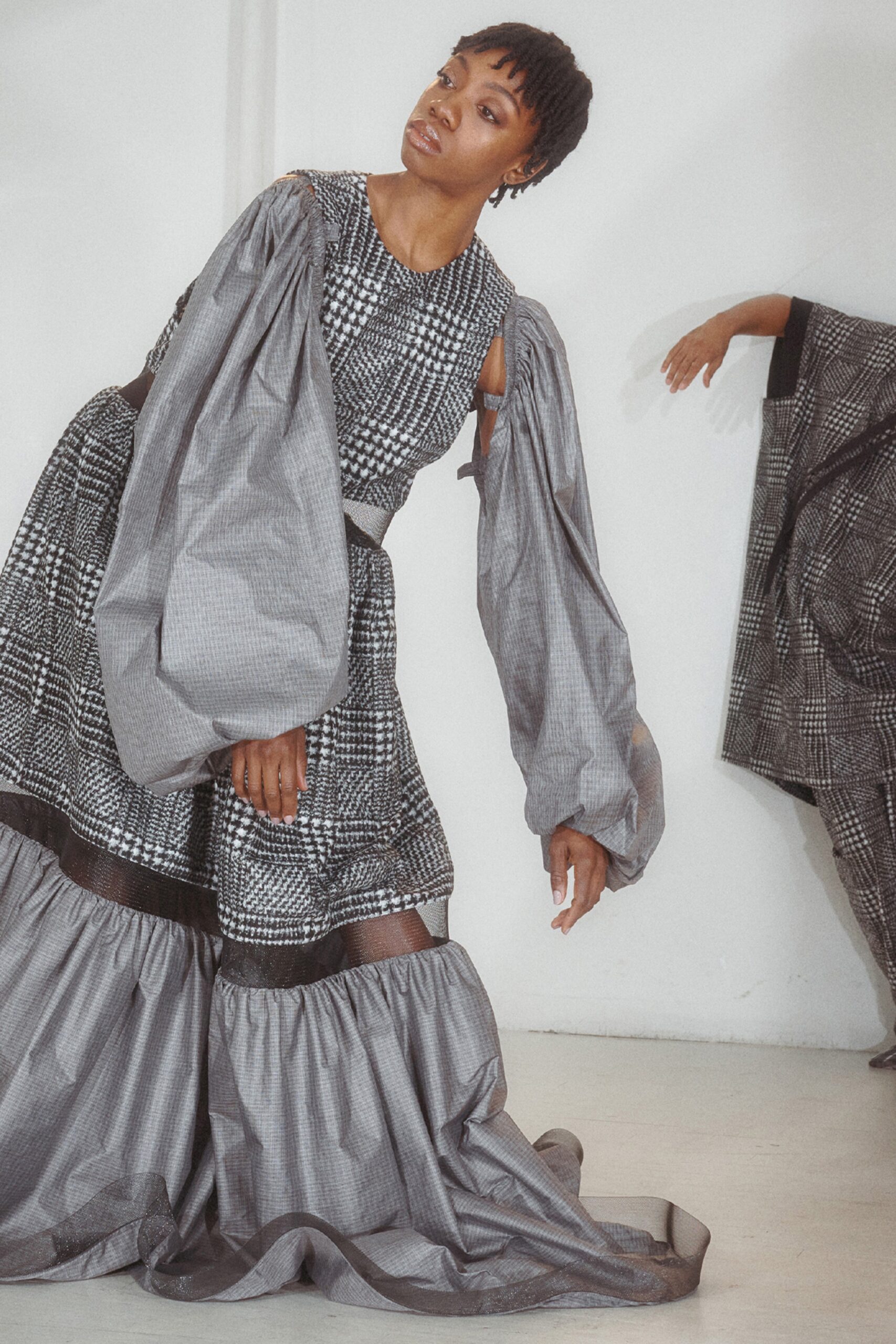 This dress features a relaxed silhouette with dramatic long sleeves and a high neckline, creating a striking visual impact. The tiered design and pleated details add depth and movement, enhancing the overall aesthetic with a contemporary flair.