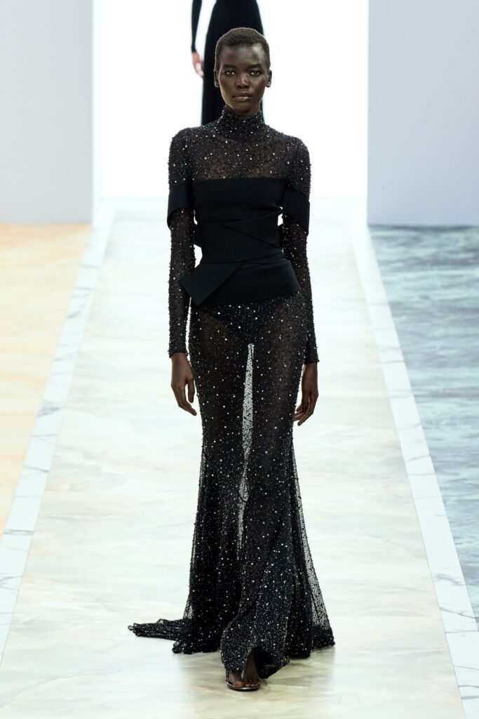 A fitted black sheer dress with long sleeves, a high neckline, and sequined details featuring a slit.
