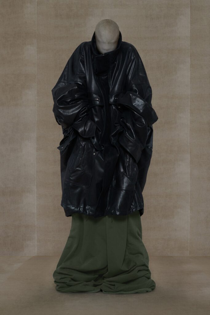 An oversized black leather coat paired with relaxed green pants, featuring a high-neck design.