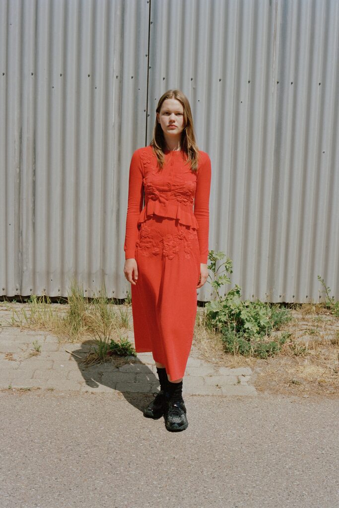 A fitted red cotton dress with long sleeves, high neckline, and layered pleated details.