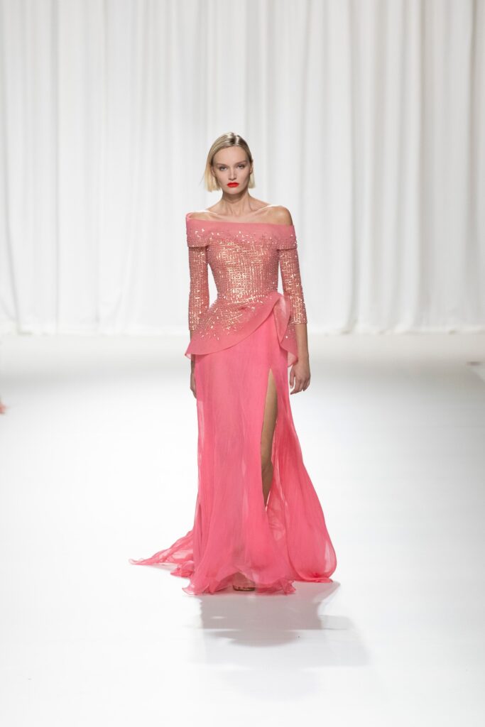 A fitted pink silk dress with an off-shoulder design, long sleeves, and a layered silhouette featuring a slit.