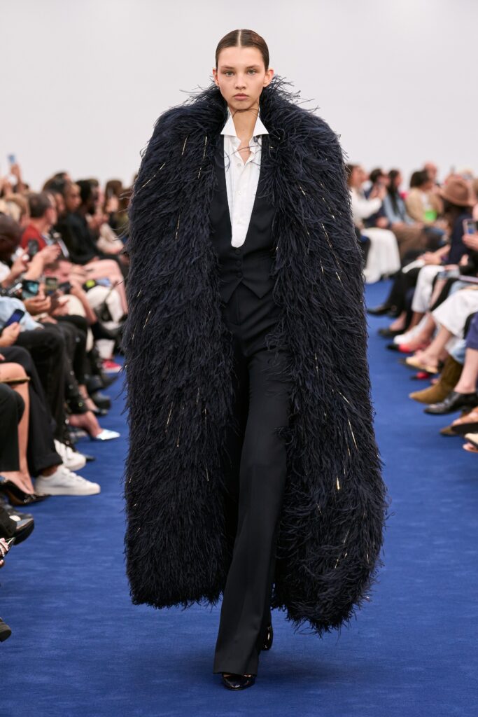 A structured navy fur coat worn over a black suit with a collared shirt.
