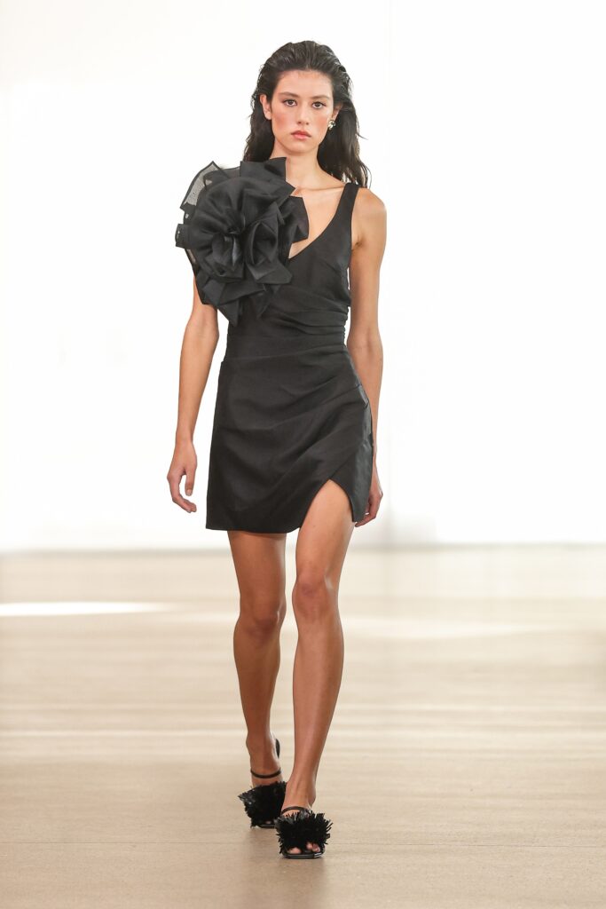 A fitted black sleeveless dress with a V-neckline and an asymmetric silhouette featuring a slit.