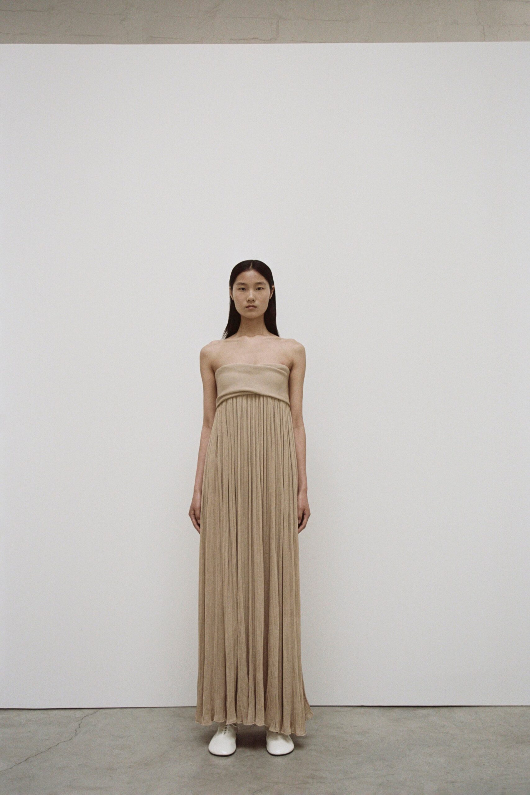 A fitted beige silk sleeveless maxi-dress with a high neckline and pleated design.