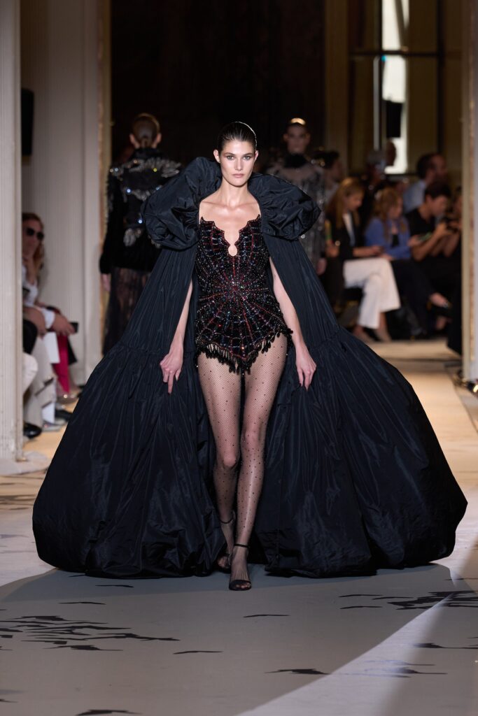 A fitted black silk dress with a V-neckline, layered design, and a dramatic cape.