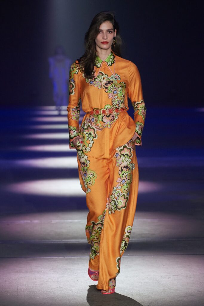 A relaxed orange silk jumpsuit with long sleeves, a collared neckline, and vibrant printed details.